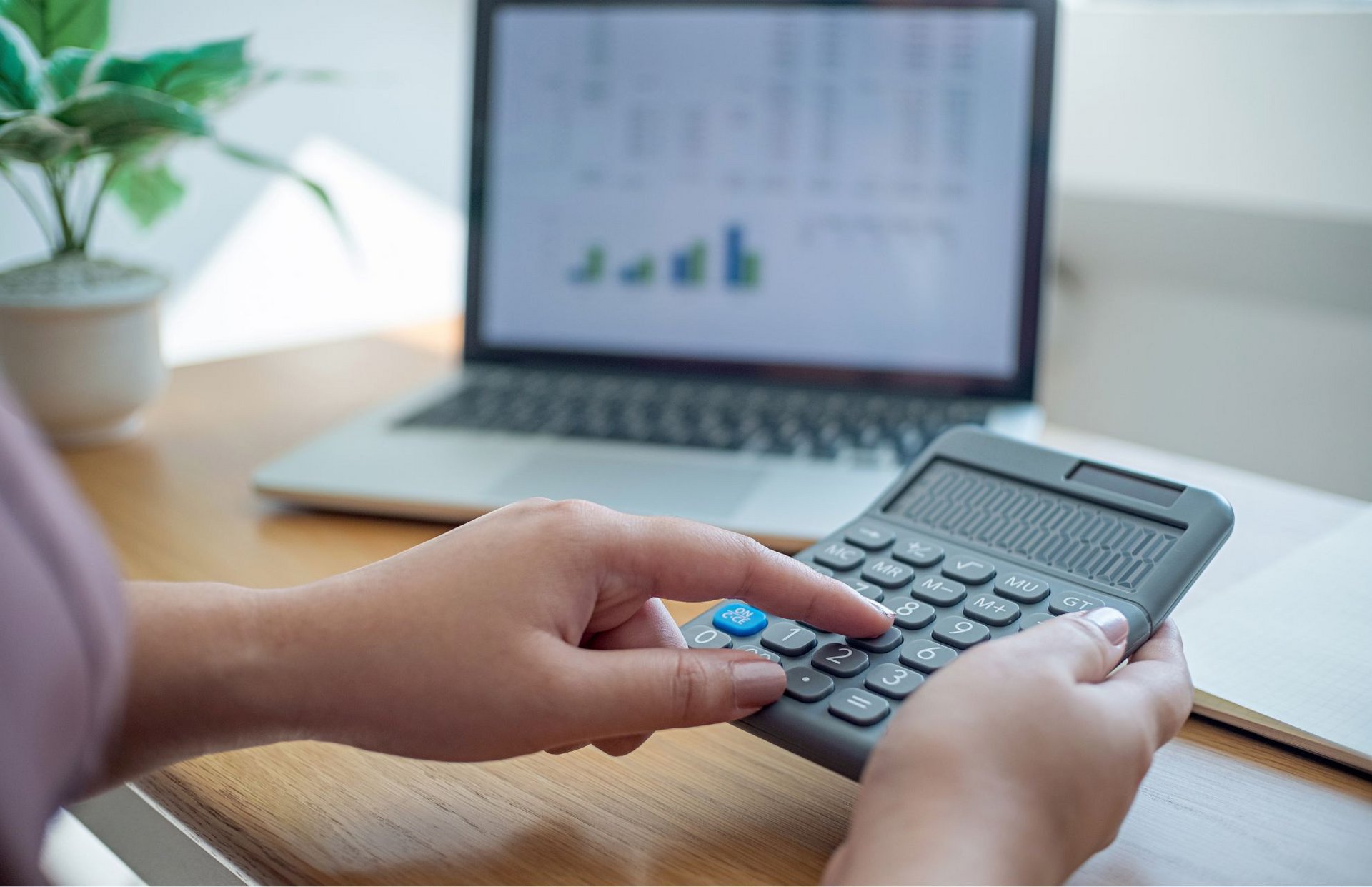 hand mit Taschenrechner, die Zahlen eintippt, inm Hintergrund ein Laptop mit einer Rechung auf dem Bildschirm
